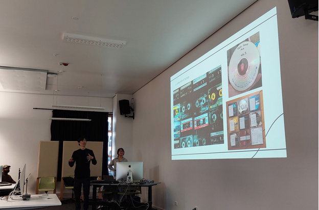 Jon explaining someting in front of a classroom. There is someone assisting with the presentation. Projected on the class wall are images of cassette tapes, presumably mixtapes.