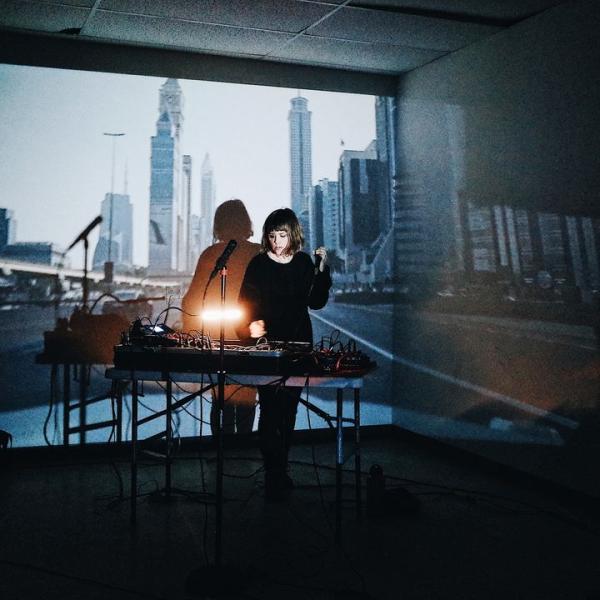 Jess Playing live, an array of electronics in front of her, and a city scene projected on the wall behind her.