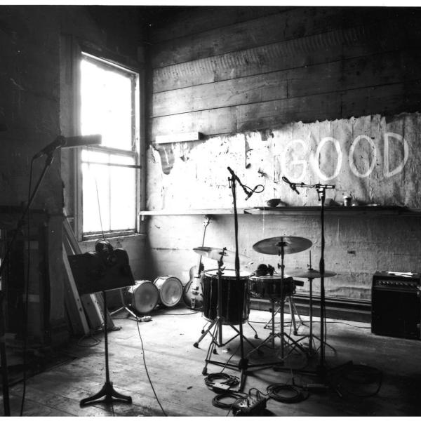 ARC Building Rehearsal Room, black and white photograph of a rehearsal room with one window beaming in bright light into an otherwise very dark room. A drumset, some amps, a music stand and a few mirrors are the only objects in this sparse dingey space.  