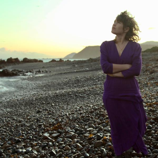 Jess Stands on a rocky beach as the sun is setting. She looks to the sky and has her arms crossed.