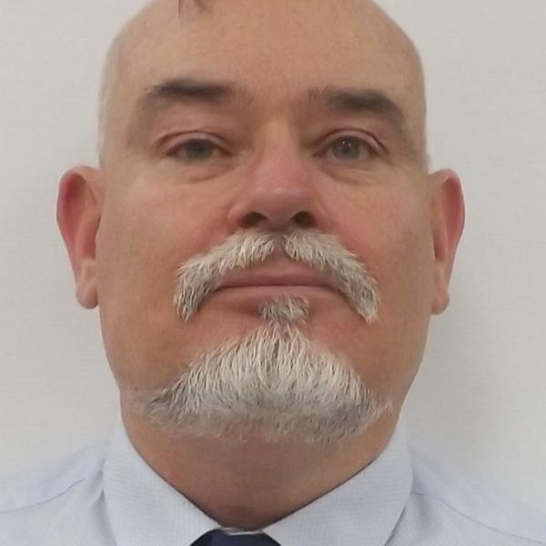 a portrait of Sam. He is looking directly at the camera with a strong and clear face. He has a well groomed goaty and moustache. He is wearing a shirt and tie.