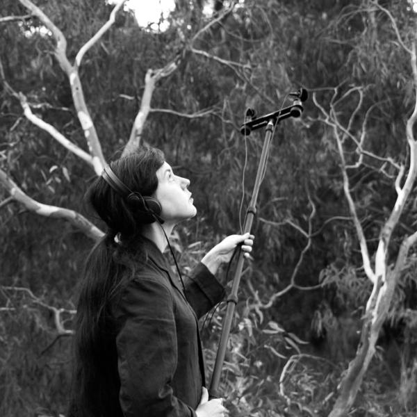 Sally in the bush. She is wearing headphones and has field recording mics on the end of a large boom