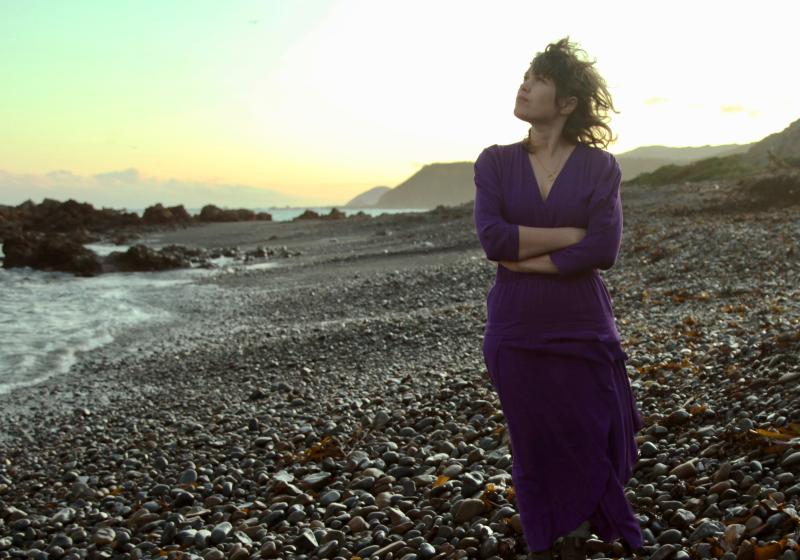 Jess stands on a rocky beach at sunset. She looks to the sky and her arms are folded