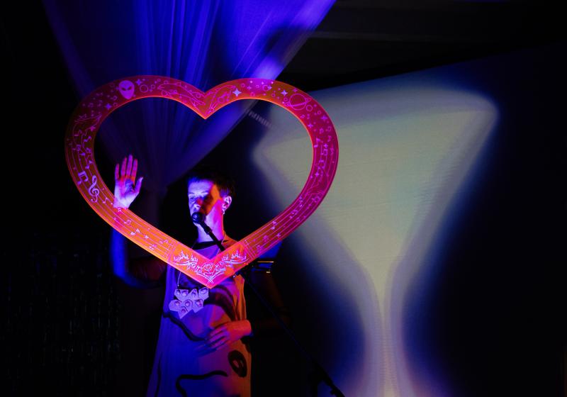 Gemma Is looking through a love heart sculpture, holding her hand up in and open gesture.