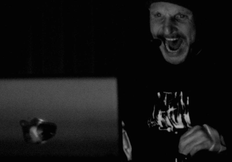 Drew is at a laptop, his mouth wide open in projection of sound. His face is scrunched in power. He has a headset microphone on.