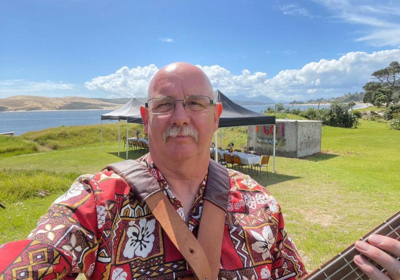 Derek is outside in the sunshine. He is holding a bass. He is bald with grey hair on the edges. He is wearing glasses. The is a gazebo behind him with tables and chairs set up underneath