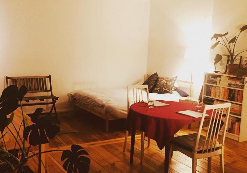 A very tidy bedroom. A table with a red table cloth. There are two glasses of water set on the table, two pads of paper, and two chairs.