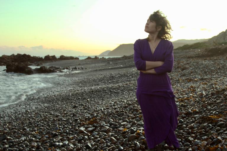 Jess Stands on a rocky beach as the sun is setting. She looks to the sky and has her arms crossed.